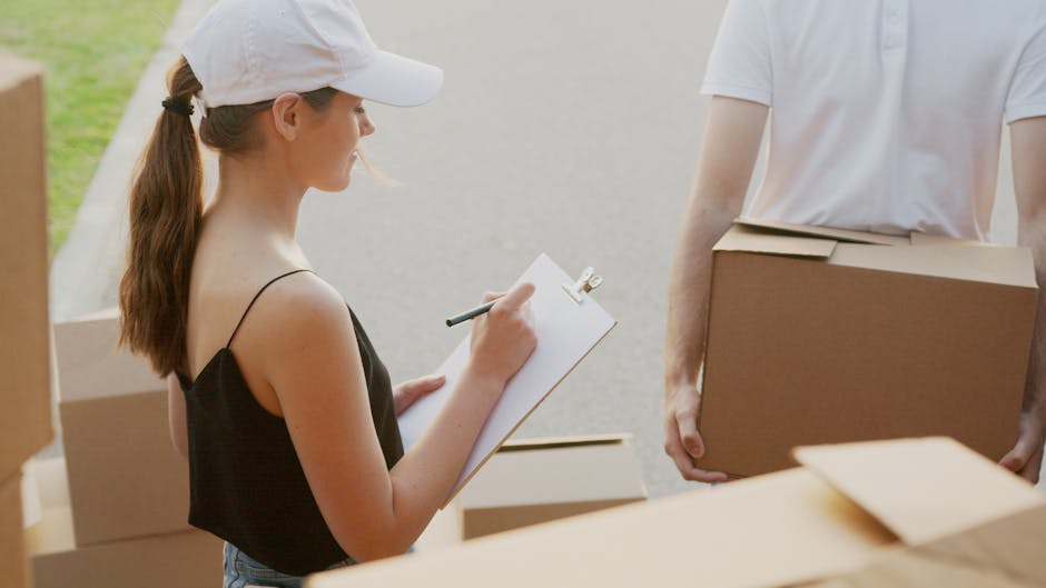 A woman with long brown hair wearing a white cap and black spaghetti strap top is outdoors, holding a clipboard and pen while taking notes during a house removal process. She is standing next to several cardboard boxes, some open and others sealed with packing tape, which are stacked on a pavement near a property entrance. In front of her, a man wearing a white t-shirt holds a medium-sized cardboard box, preparing for loading onto a vehicle. Visible in the background is a driveway or street, with natural daylight illuminating the scene. The setting appears to be part of a home relocation, with ongoing packing and furniture transport activities. The image suggests careful planning characteristic of professional removals, such as those provided by Belgravia Man and Van, and highlights packing, handling, and loading elements typical in moving services around the home.