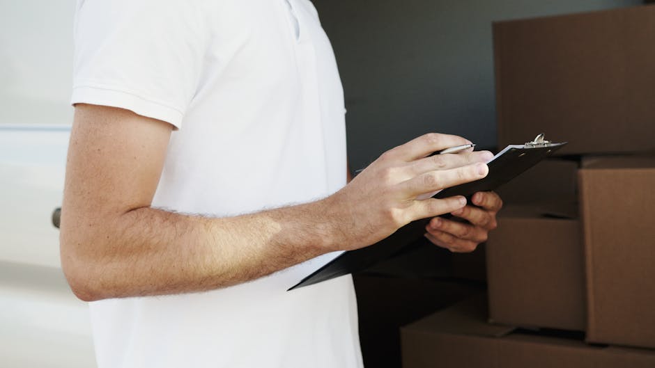 Close-up of a person in a white t-shirt holding a black clipboard and pen, standing in front of stacked cardboard moving boxes in a home environment during a house removal. The individual appears to be recording or reviewing packing details as part of a furniture transport or packing and moving process. The boxes are made of brown cardboard with some wrapped in protective plastic, placed on a wooden surface, possibly inside a corridor or a storage area during the loading process. This scene is captured under indoor lighting, highlighting the preparations and logistical aspects involved in home relocation, with a focus on domestic moving preparations. Belgravia Man and Van is involved in such removals services, supporting clients with efficient packing and transportation solutions.