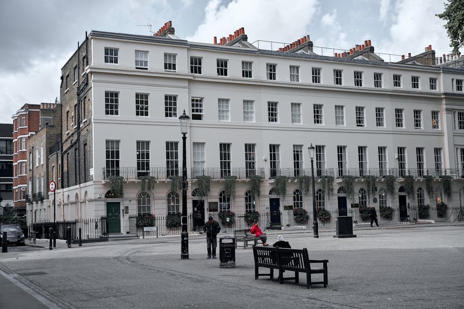 A street scene in Belgravia featuring a large white multi-story building with multiple evenly spaced sash windows and decorative greenery on the ground floor. Several black lampposts line the pavement
