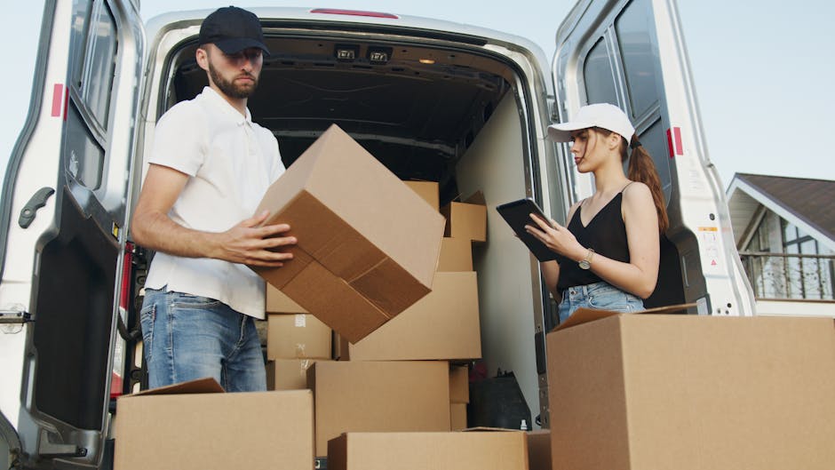 A man and a woman are engaged in the home relocation process at the back of a white moving van, which is parked outside a residential property during daylight. The man, wearing a dark baseball cap, white polo shirt, and blue jeans, is holding a large cardboard box, preparing to load or unload it from the van. Inside the van are several neatly stacked cardboard boxes of various sizes, some wrapped in plastic or packing materials. The woman, wearing a white cap, black sleeveless top, and denim shorts, is standing nearby, focused on a tablet device, possibly coordinating or confirming the packing and moving process. The rear doors of the van are open, revealing a spacious interior with additional boxes ready for transport. The environment appears to be an outdoor driveway or street, with a modern house visible in the background. This scene illustrates professional furniture transport, packing, and loading activities typical during a house move, with [COMPANY_NAME] providing targeted house removal services in the Belgravia area, supporting seamless and secure home relocations.