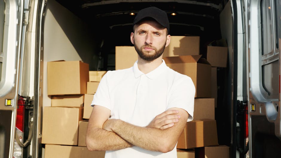 A young man with a beard, wearing a black cap and a white polo shirt, stands with arms crossed inside the open rear of a moving van loaded with multiple cardboard boxes. The boxes, varying in size and securely sealed with packing tape, are stacked neatly against the van's interior walls. Some boxes are wrapped with protective plastic wrap, indicating careful packing for a house relocation. The van is parked on a residential street, with the slightly blurred exterior showing a pavement and possibly a glimpse of nearby buildings. Belgravia Man and Van's vehicle is prepared for furniture transport and moving logistics, supporting a professional packing and moving service. The lighting inside the van is bright, highlighting the packaging materials and the person's focused expression, reflecting readiness for a delicate move in conjunction with their house removals service linked to Eaton Square in Belgravia.