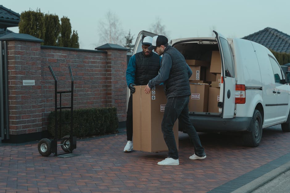 A man and a woman are engaged in the home relocation process at the back of a white moving van, which is parked outside a residential property during daylight. The man, wearing a dark baseball cap, white polo shirt, and blue jeans, is holding a large cardboard box, preparing to load or unload it from the van. Inside the van are several neatly stacked cardboard boxes of various sizes, some wrapped in plastic or packing materials. The woman, wearing a white cap, black sleeveless top, and denim shorts, is standing nearby, focused on a tablet device, possibly coordinating or confirming the packing and moving process. The rear doors of the van are open, revealing a spacious interior with additional boxes ready for transport. The environment appears to be an outdoor driveway or street, with a modern house visible in the background. This scene illustrates professional furniture transport, packing, and loading activities typical during a house move, with [COMPANY_NAME] providing targeted house removal services in the Belgravia area, supporting seamless and secure home relocations.